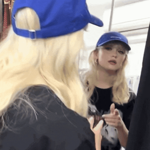 a blonde woman wearing a blue hat is pointing at herself in a mirror