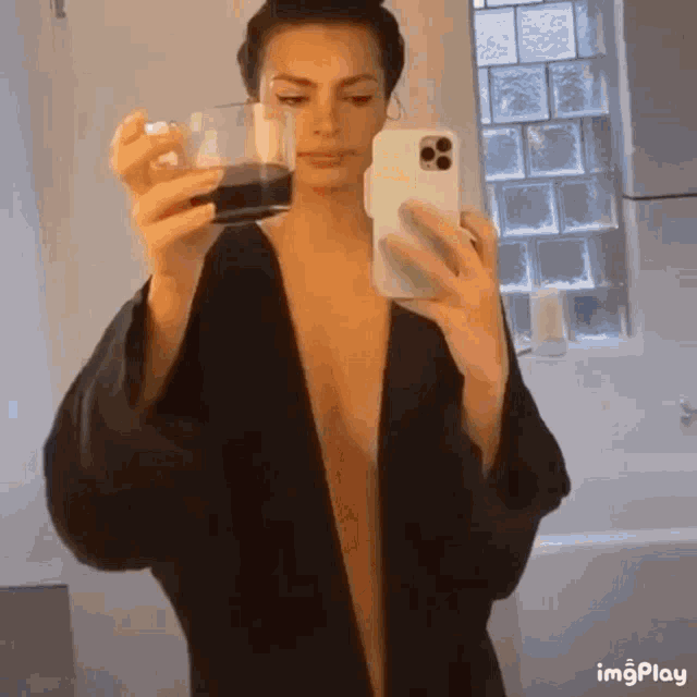 a woman in a robe is taking a picture of herself in a mirror while holding a glass of coffee .