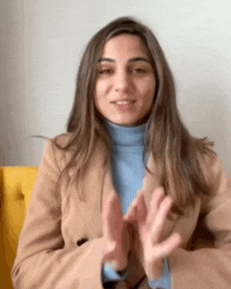 a woman in a tan coat and blue turtleneck is clapping her hands while sitting on a yellow chair .