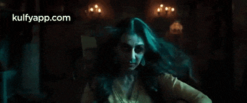 a woman in a yellow shirt is standing in a dark room with the website kulfyapp.com in the corner .
