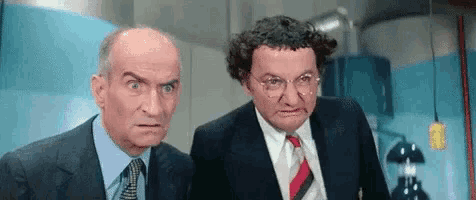 two men in suits and ties are standing next to each other in a room and making funny faces .