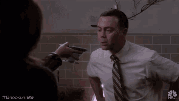 a man in a white shirt and brown tie is talking to a woman in a bathroom with #brooklyn99 written on the bottom