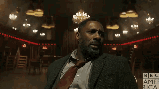 a man in a suit and tie sits in a dark room with bbc america written on the bottom right