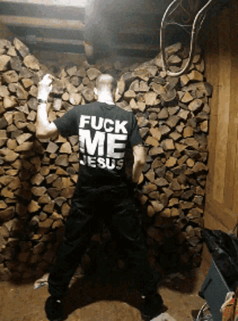 a man wearing a black shirt that says fuck me jesus is standing in front of a pile of logs