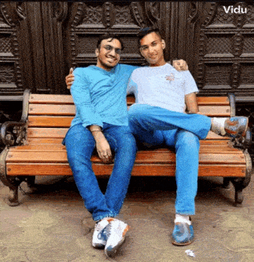 two men are sitting on a wooden bench and one has his arm around the other 's neck