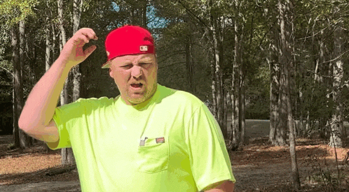 a man in a yellow shirt and a red hat is standing in the woods .