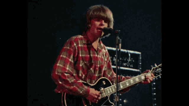 a man in a plaid shirt plays a guitar and sings into a microphone