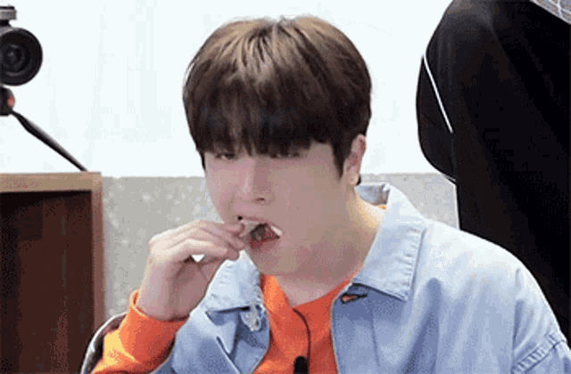 a young man in a denim jacket and orange sweater is eating a piece of food .
