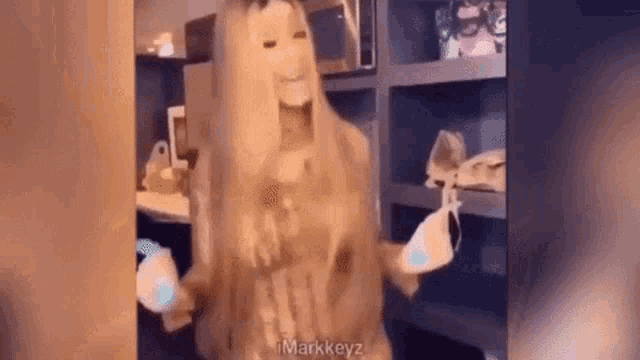 a woman with long blonde hair is standing in front of a shelf holding a pair of glasses .