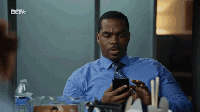 a man in a blue shirt and tie is looking at his cell phone with a bet logo behind him