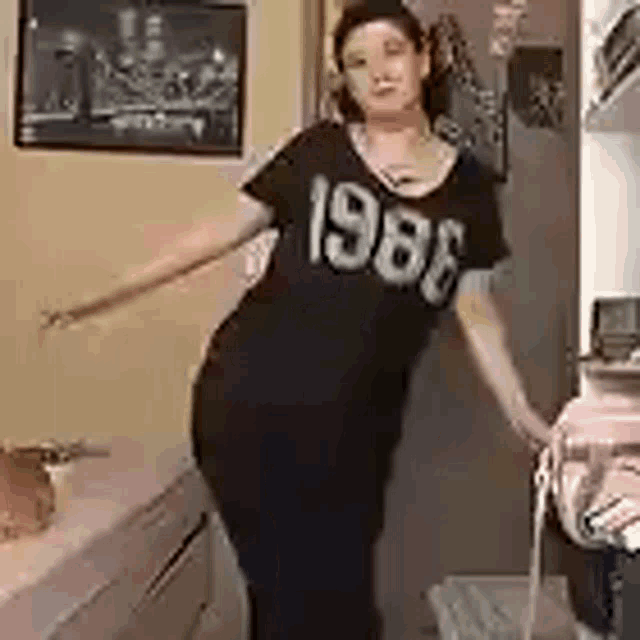 a woman is standing in a kitchen wearing a black shirt with the year 1988 on it .