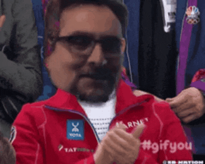 a man wearing glasses and a red jacket is giving a thumbs up while sitting in a crowd .