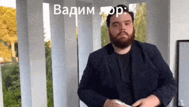 a man with a beard is standing in front of a white pillar holding a cell phone in his hand .
