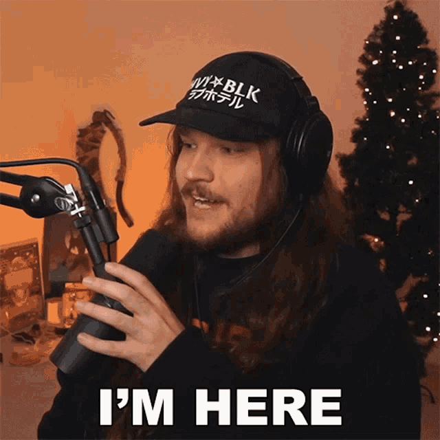 a man wearing headphones and a hat says " i 'm here " in front of a christmas tree