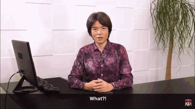 a man in a purple shirt is sitting at a desk with his hands folded and says what