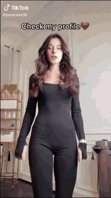 a woman in a black jumpsuit is standing in front of a wall with a tiktok caption that says check my profile