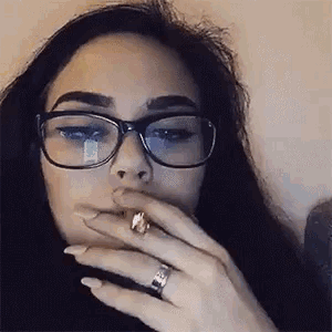 a woman wearing glasses is smoking a cigarette while wearing a ring on her finger .