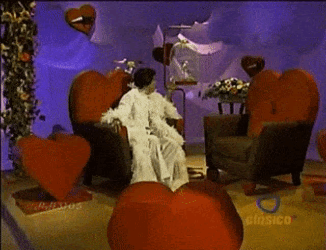 a woman in a white robe sits in a chair in a room with red hearts and the word clasico on the bottom