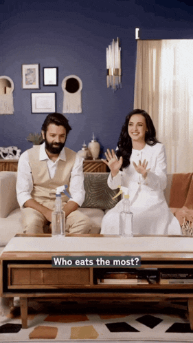 a man and a woman are sitting on a couch with a table that says who eats the most