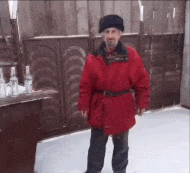 a man in a red jacket and black hat is standing in front of a wooden fence holding a bottle of vodka .