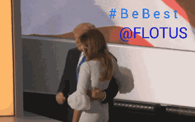 a man in a suit and tie is hugging a woman in a white dress in front of a sign that says be best @flotus