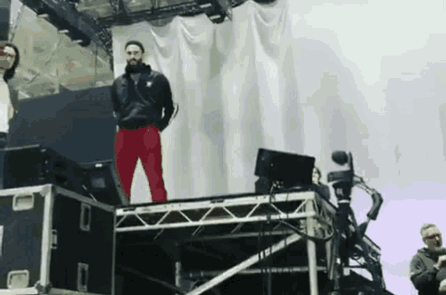 a man in red pants stands on a stage with a white curtain behind him