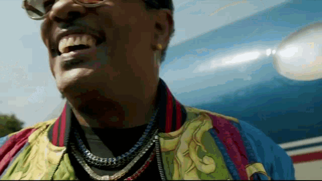 a man wearing a colorful jacket and necklaces is smiling in front of an airplane