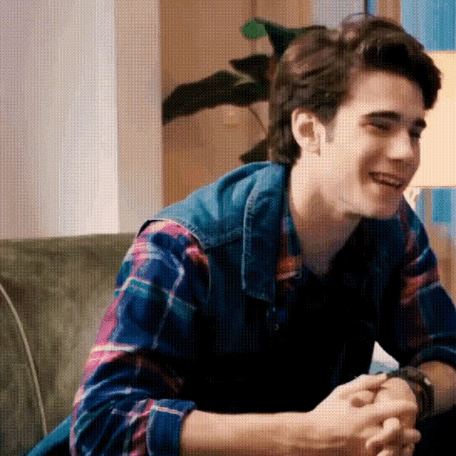 a young man is sitting on a couch wearing a plaid shirt and a denim vest and smiling .