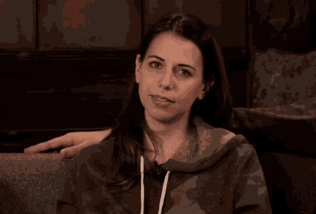 a woman wearing a camo sweatshirt winks while sitting on a couch