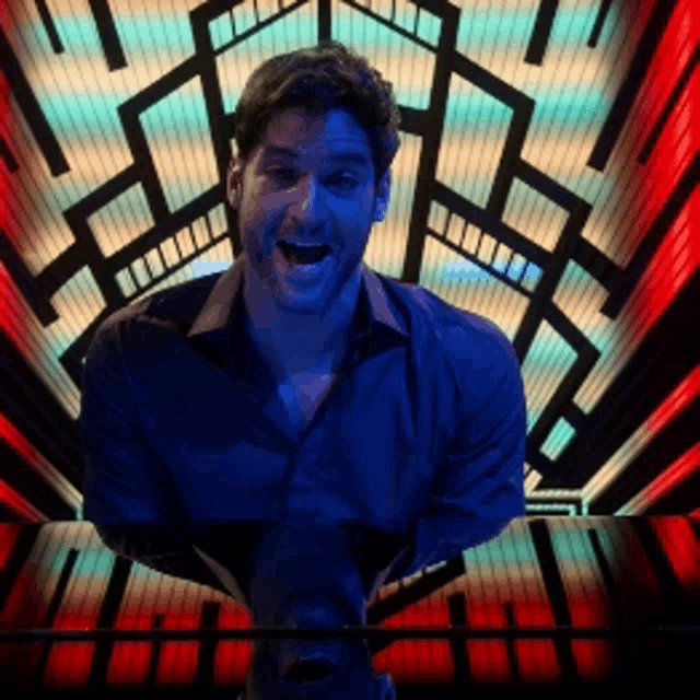 a man in a blue shirt stands in front of a colorful background with his mouth open