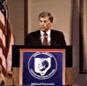 a man in a suit and tie stands at a podium with a shield on it that says cumberland university