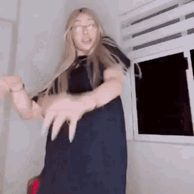 a woman in a black dress and glasses is dancing in a room in front of a window .