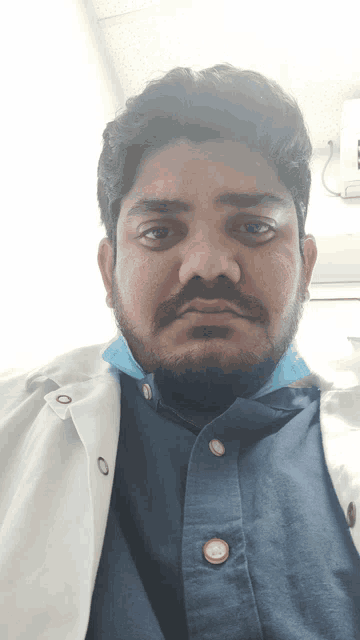 a man with a beard wearing a blue shirt and a white coat looks at the camera