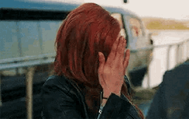 a woman with red hair is covering her face with her hands in front of a car .
