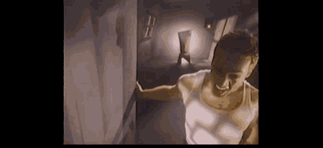 a man in a white tank top is standing in a dark room with a chair in the background .