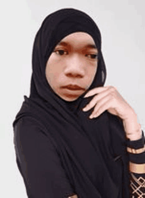 a woman wearing a black hijab and a black shirt is standing with her hand on her chin .