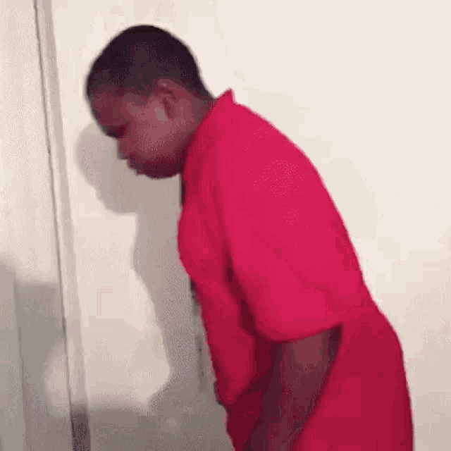 a man in a red shirt is leaning against a wall and looking down .