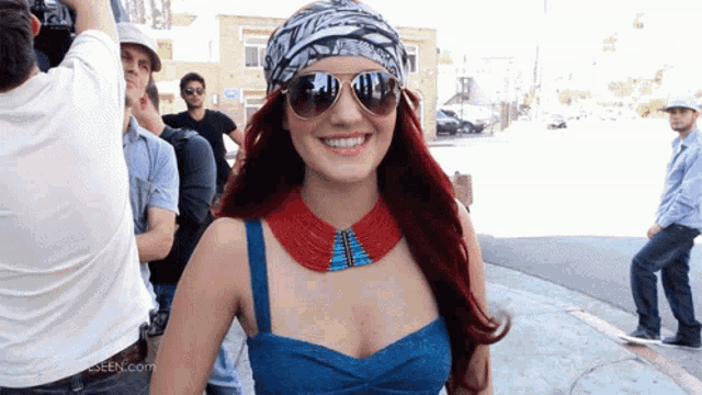 a woman wearing sunglasses and a bandana smiles in front of a sign that says ' seen.com '