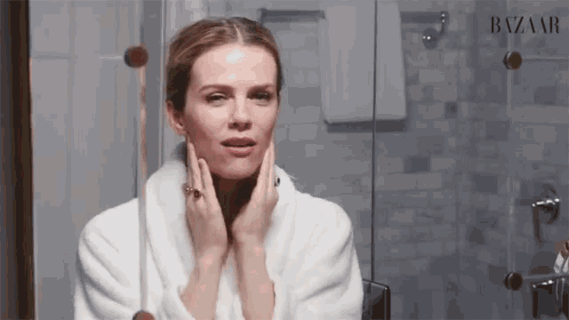 a woman in a bathrobe looks at her face in front of a mirror with the word bazaar on it
