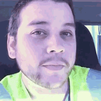 a man with a beard and a yellow vest is sitting in a car and looking at the camera .