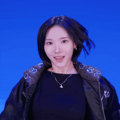 a woman wearing a black jacket and a necklace is standing in front of a blue wall
