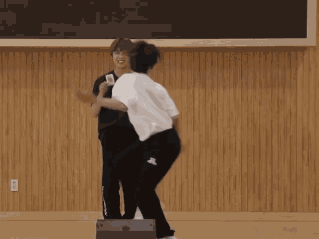 two young men are dancing in front of a wooden wall and one of them has a black shirt with a blue bear on it