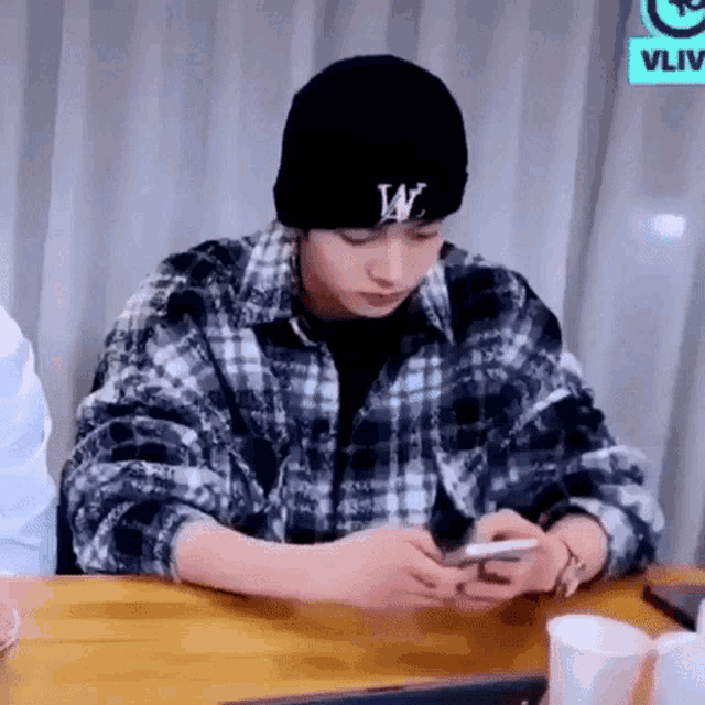 a young man wearing a plaid shirt and a black beanie is sitting at a table looking at his cell phone .