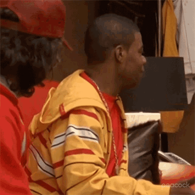 a man wearing a yellow and red striped jacket is sitting next to another man wearing a red shirt .