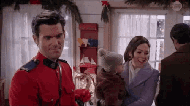 a man in a red uniform is holding a baby while a woman in a purple coat looks on .