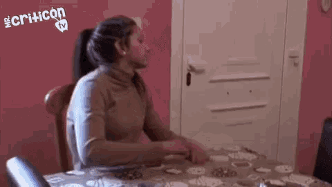 a woman sits at a table in front of a door with criticon tv in the corner