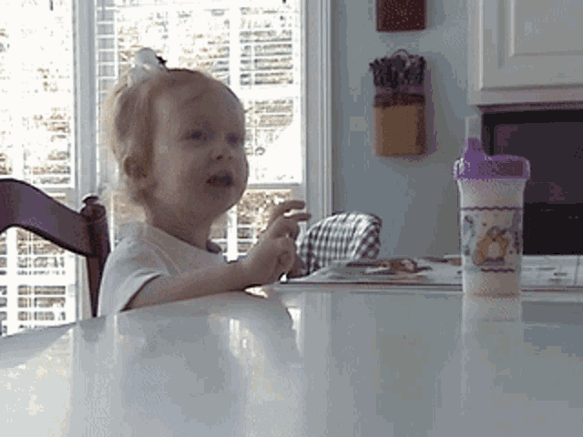 a little girl sits at a table with a purple cup