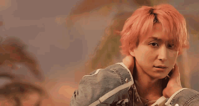 a young man with pink hair is wearing a denim jacket and holding his neck .