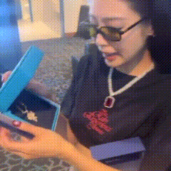 a woman wearing sunglasses is opening a blue box with a necklace in it .