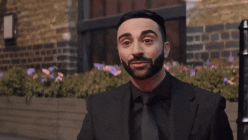 a man with a beard wearing a black suit and tie is smiling with his eyes closed .